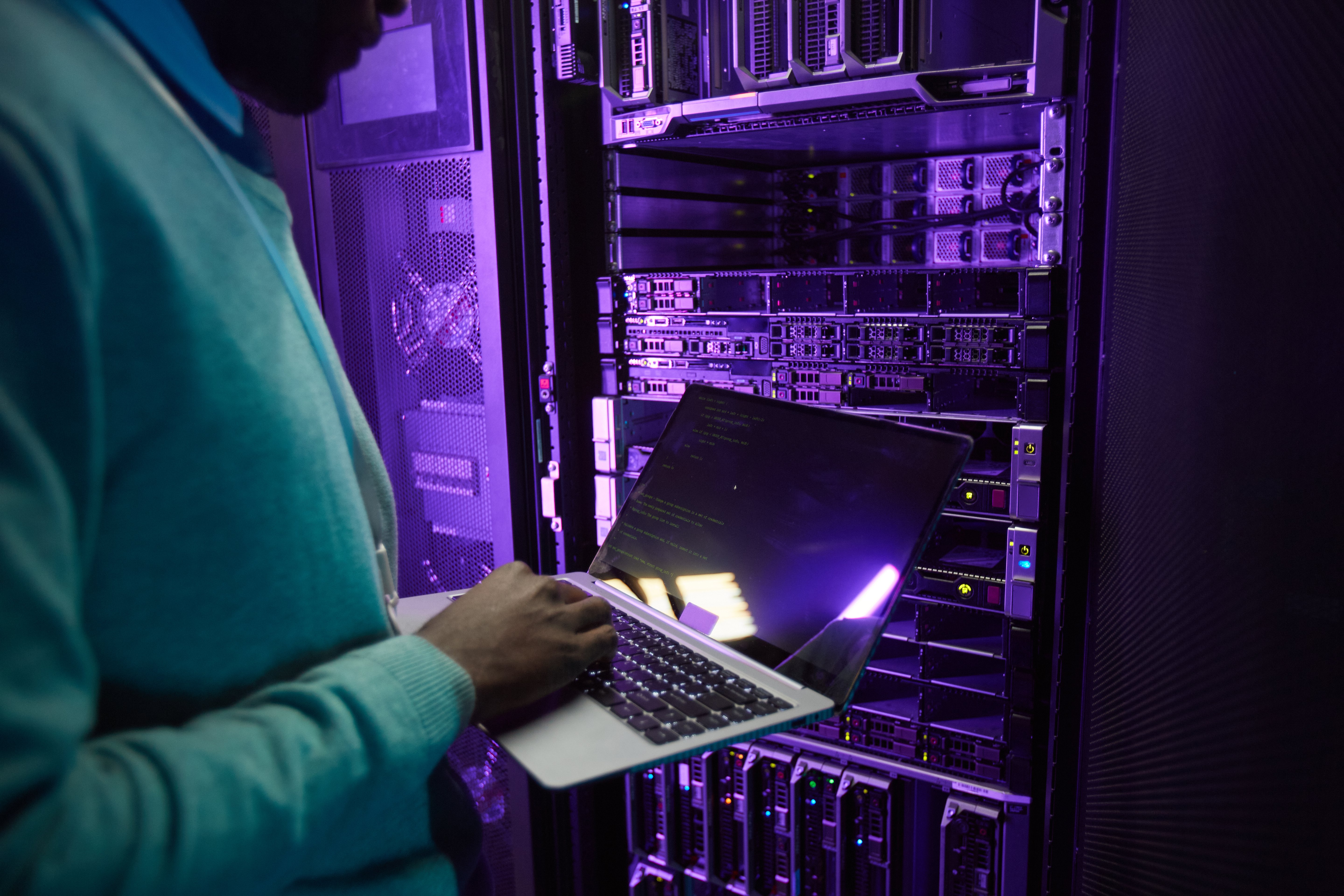 cropped-shot-african-american-data-engineer-holding-laptop-while-working-with-supercomputer-server-room-lit-by-blue-light-copy-space